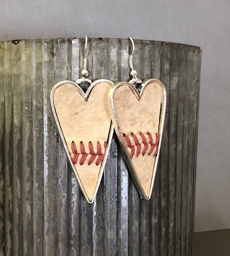Puede incluir: Pendientes en forma de coraz&oacute;n con tem&aacute;tica de b&eacute;isbol. Los pendientes presentan un fondo beige claro con costuras rojas, enmarcados por un borde de metal plateado. Cuelgan de ganchos plateados, ofreciendo un accesorio &uacute;nico.