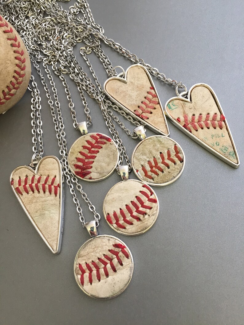 Puede incluir: Una colecci&oacute;n de collares con tem&aacute;tica de b&eacute;isbol con cadenas plateadas. Los colgantes presentan secciones de pelotas de b&eacute;isbol con costuras rojas en varias formas, incluyendo c&iacute;rculos y corazones. Un bal&oacute;n de b&eacute;isbol es visible en el fondo.