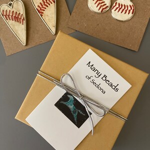Puede incluir: Pendientes con tem&aacute;tica de b&eacute;isbol y una caja de regalo. Se ven dos pares de pendientes: en forma de coraz&oacute;n y redondos, ambos con costuras de b&eacute;isbol. Tambi&eacute;n hay una caja de regalo con una cinta plateada y una etiqueta que dice "Many Beads of Sedona".