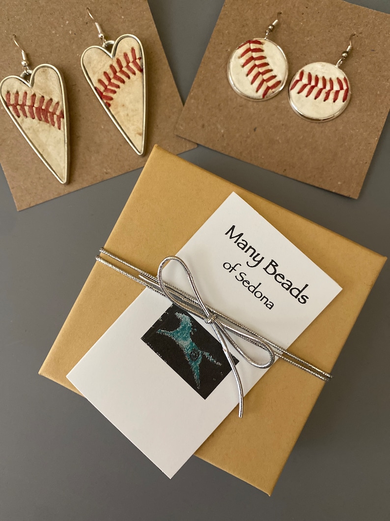 Puede incluir: Pendientes con tem&aacute;tica de b&eacute;isbol y una caja de regalo. Se ven dos pares de pendientes: en forma de coraz&oacute;n y redondos, ambos con costuras de b&eacute;isbol. Tambi&eacute;n hay una caja de regalo con una cinta plateada y una etiqueta que dice "Many Beads of Sedona".