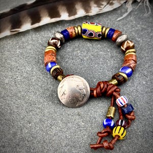 May include: A beaded bracelet featuring a silver-toned coin charm with a profile portrait, strung on brown leather cord. The bracelet includes a mix of blue, yellow, brown, and metallic beads, with a central yellow patterned glass bead.