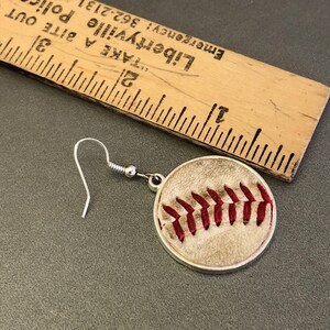 Puede incluir: Un solo pendiente con tem&aacute;tica de b&eacute;isbol con un gancho plateado. El pendiente presenta un dise&ntilde;o circular con un patr&oacute;n de b&eacute;isbol, que incluye costuras rojas sobre un fondo beige. Una regla de madera est&aacute; en el fondo.