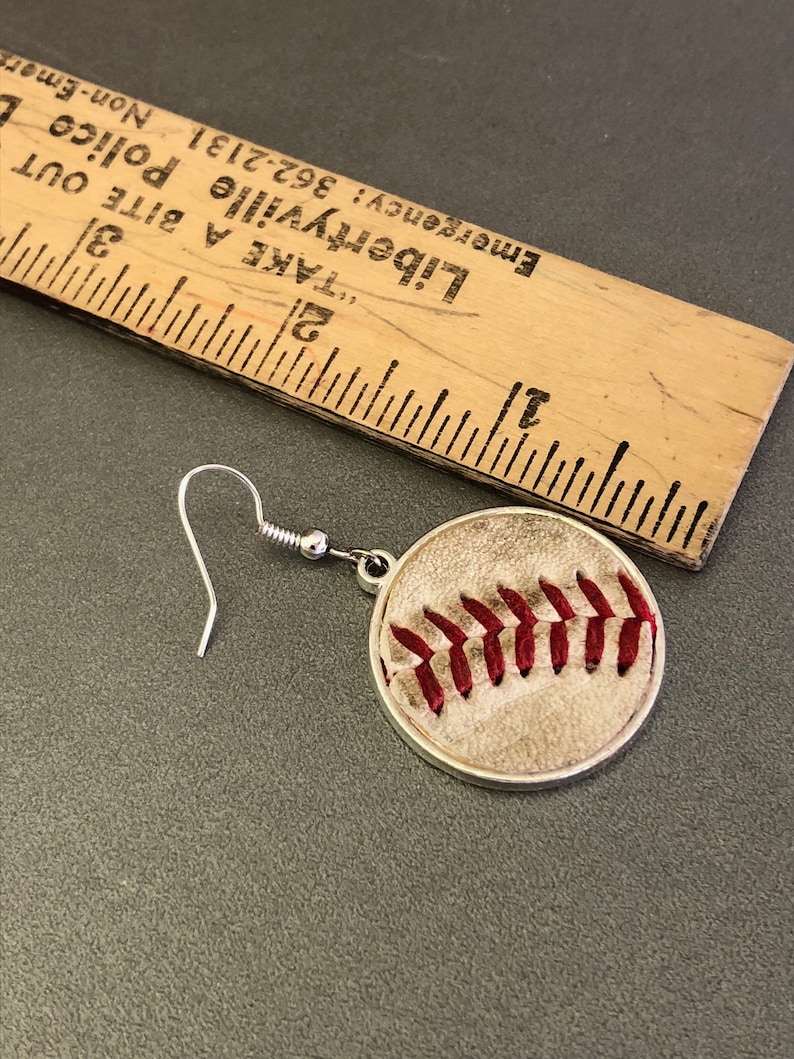 Puede incluir: Un solo pendiente con tem&aacute;tica de b&eacute;isbol con un gancho plateado. El pendiente presenta un dise&ntilde;o circular con un patr&oacute;n de b&eacute;isbol, que incluye costuras rojas sobre un fondo beige. Una regla de madera est&aacute; en el fondo.