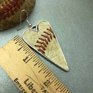 Puede incluir: Pendientes en forma de coraz&oacute;n con dise&ntilde;o de b&eacute;isbol y borde plateado. Los pendientes muestran la textura y las costuras rojas de una pelota de b&eacute;isbol. Una pelota de b&eacute;isbol y una regla de madera est&aacute;n en el fondo.