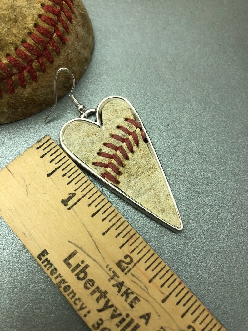 Puede incluir: Pendientes en forma de coraz&oacute;n con dise&ntilde;o de b&eacute;isbol y borde plateado. Los pendientes muestran la textura y las costuras rojas de una pelota de b&eacute;isbol. Una pelota de b&eacute;isbol y una regla de madera est&aacute;n en el fondo.