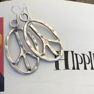 May include: Silver peace sign earrings with a hammered texture. The earrings are oval shaped and have a wire loop for hanging.