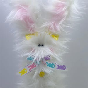 May include: A fluffy white Easter bunny decoration with pink inner ears and yellow bows. The bunny has a colorful ribbon with Easter bunny graphics and a yellow button. The bunny is a decorative item.