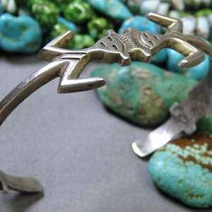 May include: A silver-toned cuff bracelet with a stylized figure design. The bracelet features an open design with a detailed figure at the center. The background includes turquoise stones in various shades of blue and green.