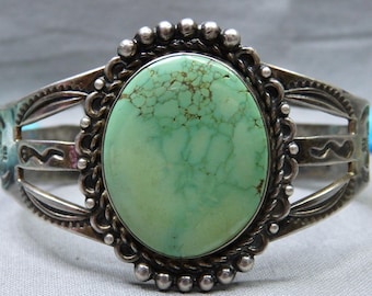 May include: A silver cuff bracelet featuring an oval, light green turquoise stone. The stone is set in a decorative bezel, surrounded by a beaded border. The bracelet has a split shank design with etched details.