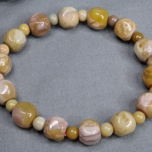 May include: A beaded bracelet featuring a repeating pattern of smooth, polished, round and cube-shaped beads in shades of mustard yellow, soft pink, and cream. The beads are strung together in a circular arrangement, showcasing a glossy finish.