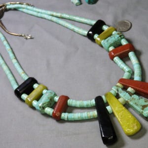 May include: A turquoise beaded necklace with black, yellow, and red accents. The necklace features a variety of stone shapes and sizes, including rectangular and teardrop-shaped pendants. The necklace has a silver clasp.