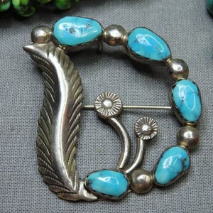 May include: A silver-toned belt buckle with a feather design on one side and a series of turquoise stones. The buckle has a decorative pin and two flower-shaped accents. The turquoise stones are oval-shaped and set in silver.