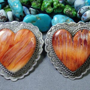 May include: Two heart-shaped pendants with orange and white patterns, set in ornate silver frames. The pendants are surrounded by turquoise and green beads, creating a striking contrast. The intricate details of the silverwork and the natural stone patterns are highlighted.