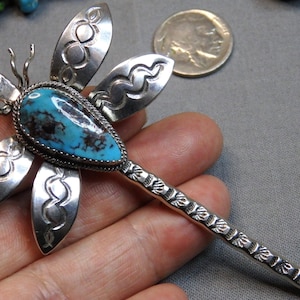 May include: A silver dragonfly brooch featuring a teardrop-shaped turquoise stone with dark matrix in the center. The four wings are decorated with stamped geometric patterns, and the long tail has a textured, segmented design.