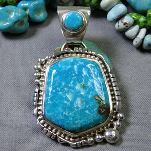 May include: A large, rectangular turquoise pendant with a silver setting. The pendant has a bright blue color with brown and white inclusions. The silver setting has a decorative border of small silver beads and a small turquoise stone at the top.