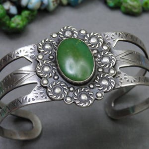May include: Silver cuff bracelet featuring an oval green stone centerpiece. The bracelet has intricate floral and geometric designs, with three parallel bands and decorative cutouts. A vintage-style piece.