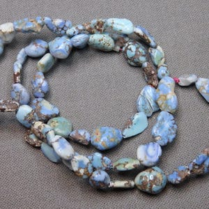 May include: A necklace composed of irregular, polished stones in shades of blue, turquoise, and brown. The stones are strung together, forming a loop. The necklace is displayed on a neutral background.