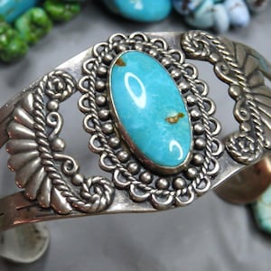May include: A silver cuff bracelet featuring a central oval turquoise stone with brown matrix inclusions. The stone is set in a scalloped bezel surrounded by a beaded border, flanked by decorative silver fan-shaped motifs and scrollwork.
