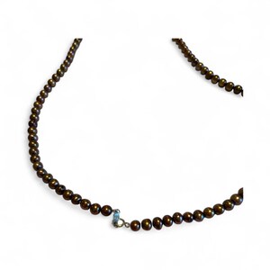 May include: A necklace composed of small, dark brown beads. The beads are strung together, forming a continuous strand. A silver-colored clasp secures the necklace. The necklace is displayed against a white background.