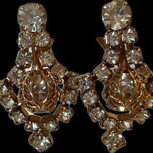 May include: A pair of vintage chandelier earrings with a gold-tone metal setting. The earrings are adorned with numerous clear, sparkling rhinestones in various sizes, creating a glamorous and eye-catching design. The earrings have a drop length of approximately 3 inches.