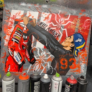 May include: A spray paint artwork on a rectangular board featuring a stylised illustration of Marc Marquez wearing a blue and yellow cap and black racing gear, holding a red racing suit. The design includes the number 93, spider motifs, and text.