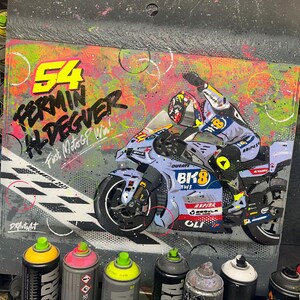 May include: A vibrant artwork featuring a motorcycle racer on a Ducati bike, with the number 54 and the name FERMIN ALDEGUER. The background is a mix of colors, with spray paint cans in the foreground. Text includes "First MotoGP Win!"