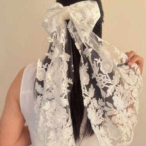 May include: White lace hair bow with a large bow in the center. The bow is made of a sheer fabric with a floral pattern.