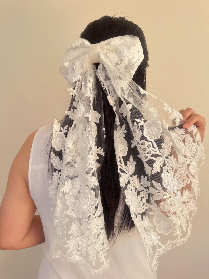 May include: White lace hair bow with a large bow in the center. The bow is made of a sheer fabric with a floral pattern.