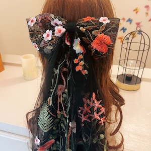 May include: Black mesh hair bow with floral embroidery in shades of red, pink, green, and blue. The bow is tied in a large, loose knot and features a flamingo design.