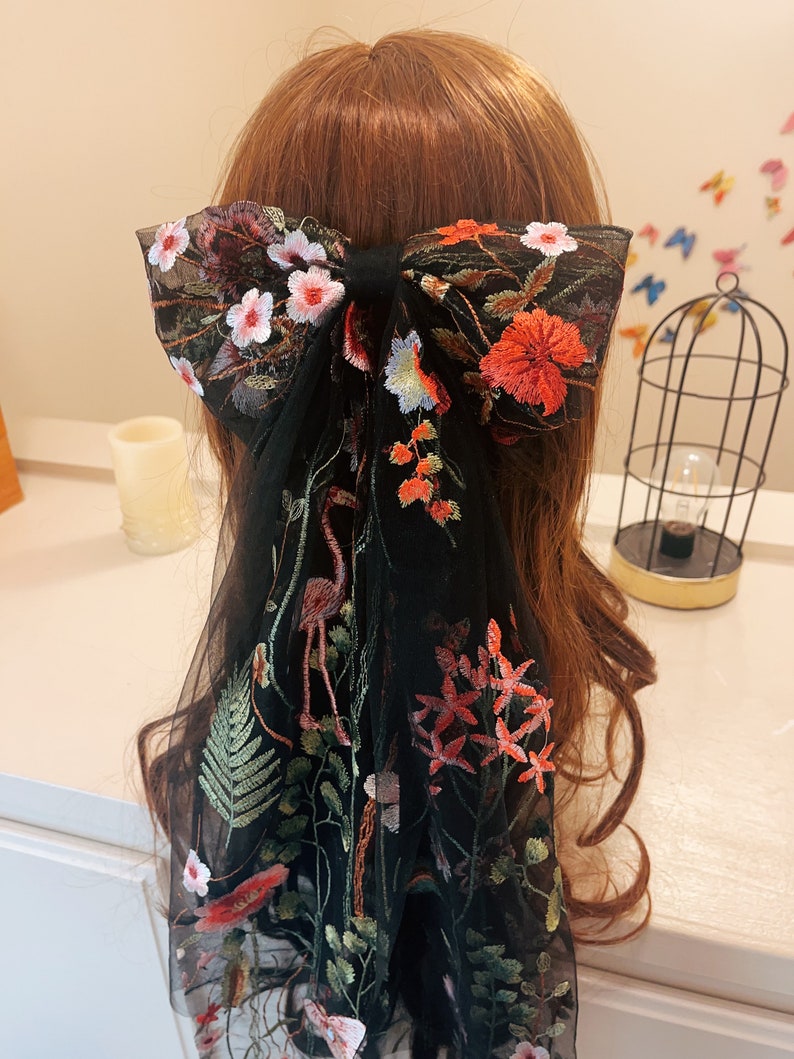 May include: Black mesh hair bow with floral embroidery in shades of red, pink, green, and blue. The bow is tied in a large, loose knot and features a flamingo design.