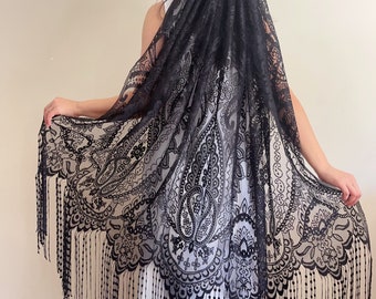 May include: Black lace veil with a paisley pattern and fringe. The veil is draped over a person's head and shoulders.