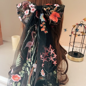 May include: Black sheer hair scarf with floral embroidery. The embroidery features pink, red, and green flowers, as well as a flamingo.