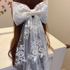 May include: A large white lace bow hair accessory with a long, flowing veil. The bow is made of delicate lace with floral patterns and a satin ribbon center. The veil also features intricate lace detailing, creating an elegant and romantic look.