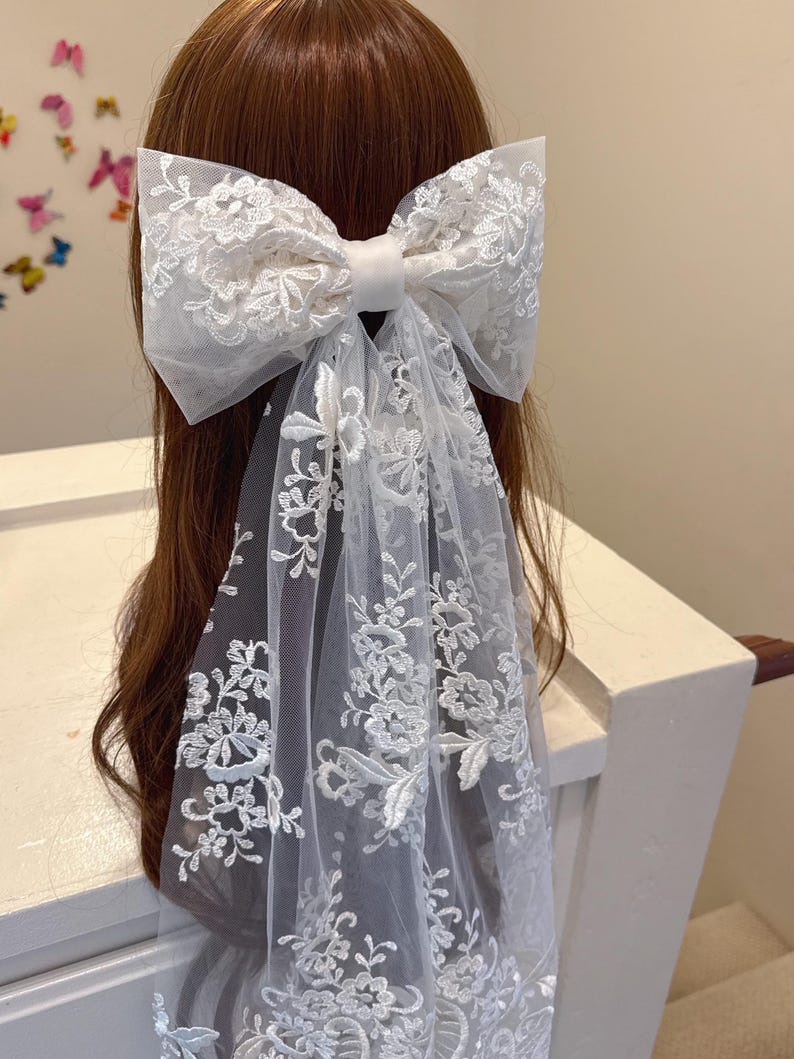 May include: A large white lace bow hair accessory with a long, flowing veil. The bow is made of delicate lace with floral patterns and a satin ribbon center. The veil also features intricate lace detailing, creating an elegant and romantic look.