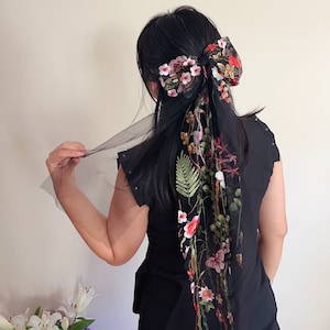 May include: A black hair bow with a large floral design in shades of pink, red, and green. The bow is made of tulle and has a long, flowing tail.