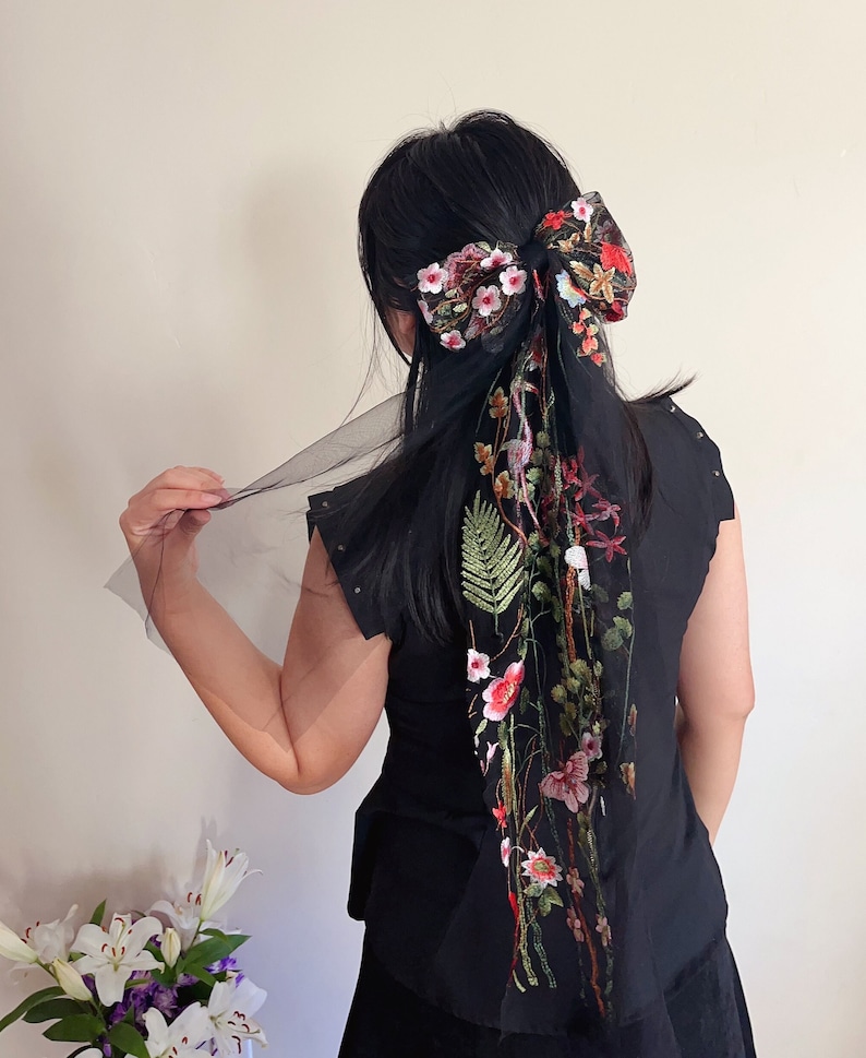 May include: A black hair bow with a large floral design in shades of pink, red, and green. The bow is made of tulle and has a long, flowing tail.