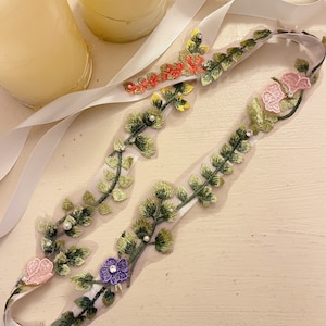 K&ouml;nnte beinhalten: Ein wei&szlig;es Satinband mit einem gr&uuml;nen, gelben, rosa und violetten floralen Stickmuster. Der Besatz ist mit kleinen perlmuttartigen Perlen und Strasssteinen verziert.