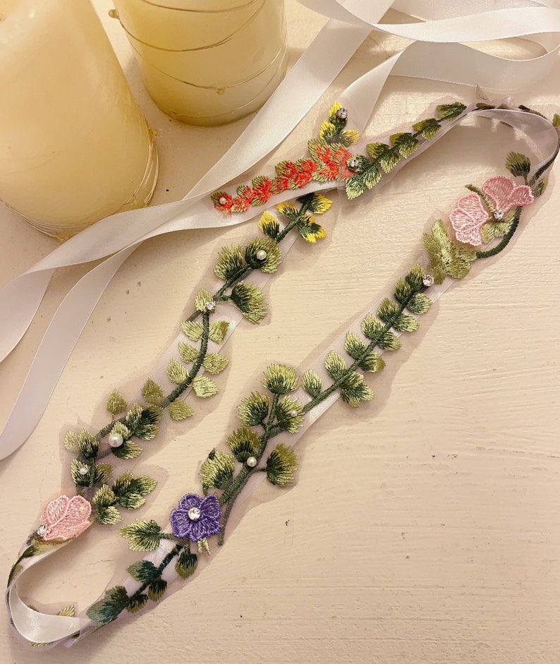 K&ouml;nnte beinhalten: Ein wei&szlig;es Satinband mit einem gr&uuml;nen, gelben, rosa und violetten floralen Stickmuster. Der Besatz ist mit kleinen perlmuttartigen Perlen und Strasssteinen verziert.