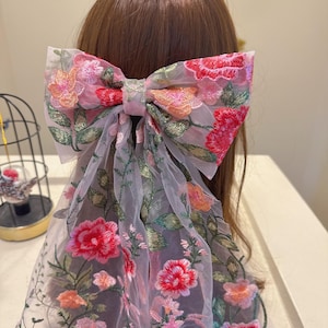 May include: A large, floral embroidered hair bow in shades of pink, red, and orange. The bow is made of sheer fabric and features detailed embroidery of flowers and green leaves. The bow is attached to a hair accessory, adding a touch of elegance.