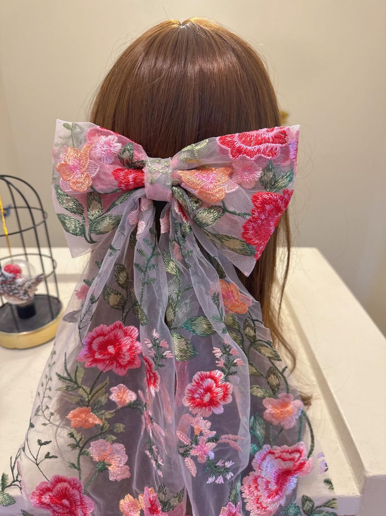 May include: A large, floral embroidered hair bow in shades of pink, red, and orange. The bow is made of sheer fabric and features detailed embroidery of flowers and green leaves. The bow is attached to a hair accessory, adding a touch of elegance.