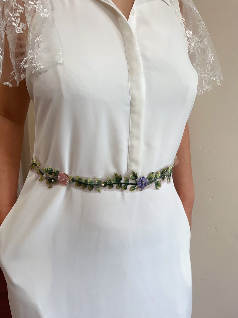 K&ouml;nnte beinhalten: Ein wei&szlig;es Kleid mit Spitzen&auml;rmeln und einem floral bestickten G&uuml;rtel.