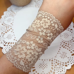 May include: A beige lace wrist cuff with a delicate floral pattern. The cuff is made of a soft, sheer fabric and has a slightly shimmery finish.