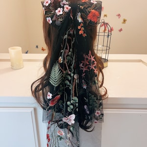 May include: Black sheer hair scarf with floral embroidery. The scarf features a large bow at the top and a long, flowing tail with delicate floral and butterfly designs.