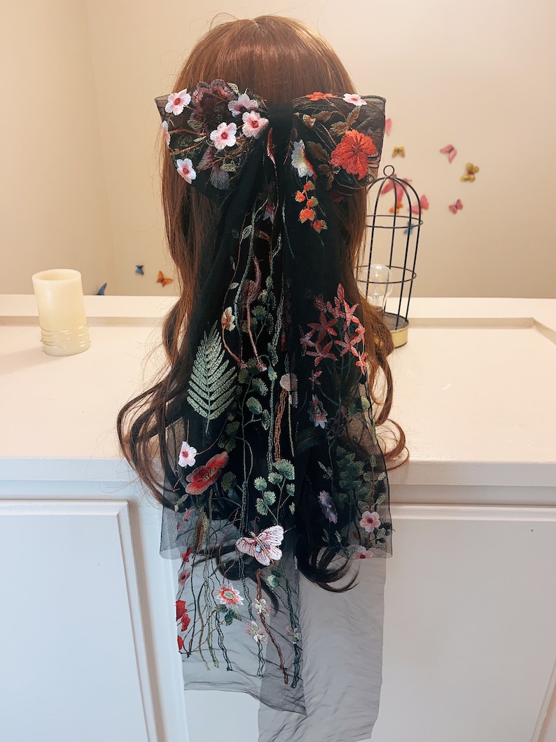 May include: Black sheer hair scarf with floral embroidery. The scarf features a large bow at the top and a long, flowing tail with delicate floral and butterfly designs.