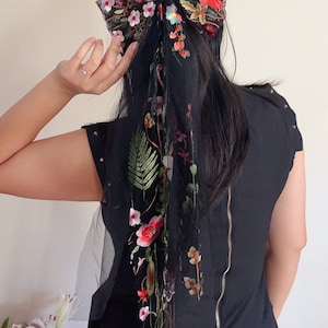 May include: A black sheer hair scarf with floral embroidery in shades of red, pink, green, and white. The scarf is tied in a bow at the top of the head and hangs down the back.