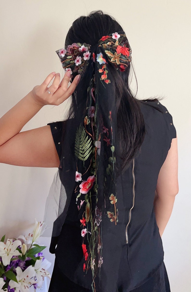 May include: A black sheer hair scarf with floral embroidery in shades of red, pink, green, and white. The scarf is tied in a bow at the top of the head and hangs down the back.