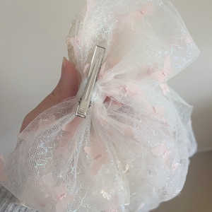May include: A white tulle hair bow with pink butterfly accents and a silver clip. The bow is made of a sheer, delicate fabric and has a sparkly finish.