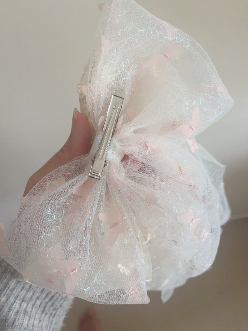 May include: A white tulle hair bow with pink butterfly accents and a silver clip. The bow is made of a sheer, delicate fabric and has a sparkly finish.