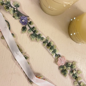 K&ouml;nnte beinhalten: Ein wei&szlig;es Band mit gr&uuml;nen und goldenen gestickten Bl&auml;ttern und rosa und violetten Bl&uuml;ten. Das Band liegt auf einer wei&szlig;en Oberfl&auml;che.