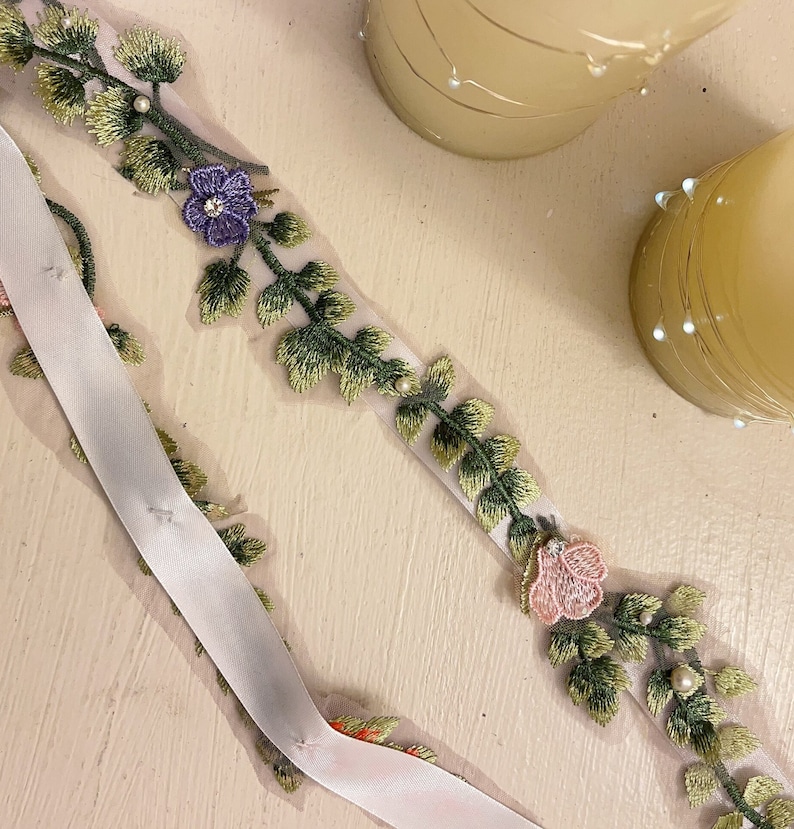 K&ouml;nnte beinhalten: Ein wei&szlig;es Band mit gr&uuml;nen und goldenen gestickten Bl&auml;ttern und rosa und violetten Bl&uuml;ten. Das Band liegt auf einer wei&szlig;en Oberfl&auml;che.